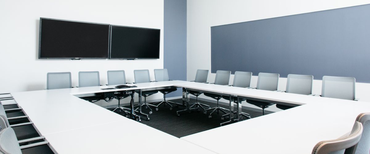 An empty boardroom with a large screen on one wall and a pin-board on the other. Four tables are organized to form a square, and surrounded with many chairs.