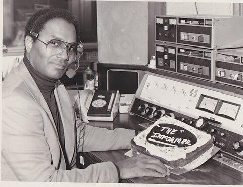 A black and white photo of FIl Fraser, Canada's first black broadcaster, sitting in a radio room. In front of him sits a cake with the words "the informer" in icing on top.