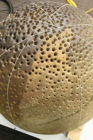 An overhead photo of the round rock with small holes. The rock is wrapped in criss-crossed string mapping out segments to be cleaned. One clean segment is a lighter brown than the rest.