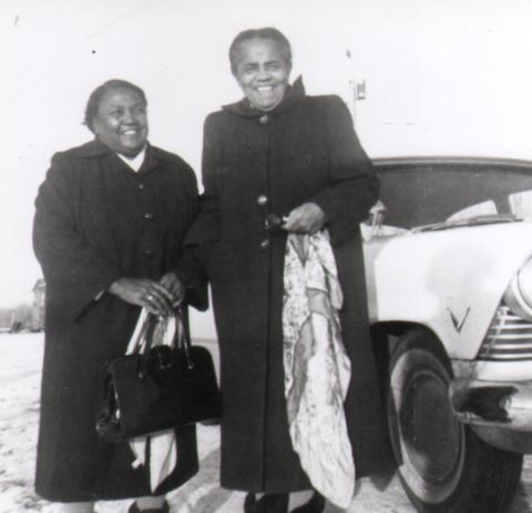 A black and white photo of two smiling women in standing in front of a car. The women's names are Lossie Lane and Ivy Beaver. The women are holding a purse and scarf.