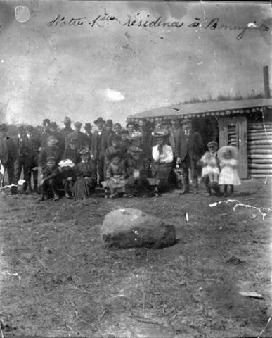 A black and white archival photo of a family standing in front of a log cabin. In the foreground a large boulder sits on the grass.