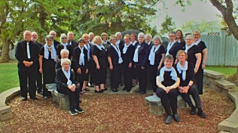 A group photo of the North Edmonton Seniors Association Choralaires. Men and women standing in a semi-circle. They are all dressed in black and white dress clothes.