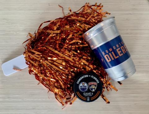 Three items lay in a pile on a wood surface: On orange metallic pom-pom, a silver cup witht the words "Edmonton Oilers", and a hockey puck with the words "Edmonton vs Florida Game 6" and the logos for the Edmonton Oilers and Florida Panthers.