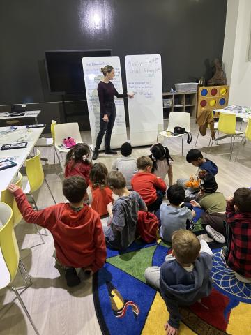 A group of students sitting cross-legged on the floor of a museum classroom. At the front of the room is a Museum staff member pointing to words written on a whiteboard.