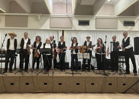 Photo of the Wild Rose Fiddlers. A group of seniors wearing black and white formal clothing. Many of them are holding fiddles, one is holding a guitar and another holds an accordian.