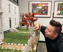 Lance Cardinal crouches down to adjust a small detail on the fence of his miniature model of St. Martin's Catholic Mission School.