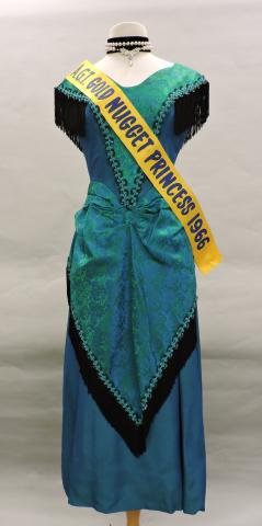 Simone Bessette's 1966 "Gold Nugget Princess" winning dress. The dress is a floor length, dark green dress with black fringe accents.