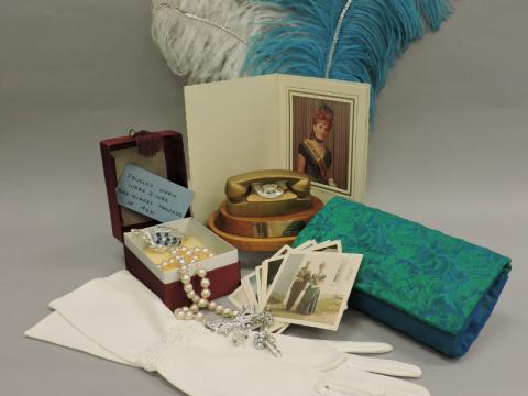 A display of personal items. White gloves, a strand of pearls, rhinestone jewellery, an open jewellery box containing a brooch and note reading "Jewelry worn when I was Gold Nugget Princess in 1966," a miniature gold telephone trophy on a wooden base, a portrait of Simone Bessette in a folded frame, several vintage snapshots, a folded turquoise patterned hand purse, and large white and turquoise feathers draped behind the display.