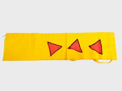 A rectangular piece of yellow cloth. There is a hem on one side, the other three sides are frayed. There are three pink triangles with black outlines painted or drawn on the right side of the cloth.