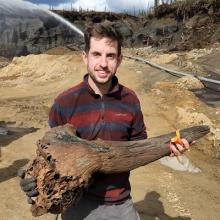 Medium shot of Scott Cocker. He is holding a large bone that has been recently excavated. He is wearing a red and black striped polo shirt and is looking at the camera with a smile on his face.