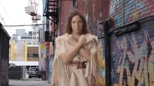 A woman in traditional clothes is standing in an urban alley. She looks at the camera with her arms crossed across her chest.