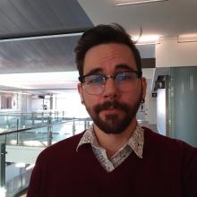 A headshot of Kyle Shanebeck. He is wearing a red sweater over a white dress shirt. He has glasses and facial hair. He is looking at the camera with a neutral face.