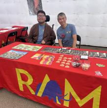 Two smiling volunteers are sitting behind a table covered in a red table cloth with the Royal Alberta Museum logo on it.