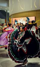 A traditional mexican dancer in a colourful dress smiles as she dances in the Royal Alberta Museum's lobby. You can see a crowd of people behind her looking on and behind them is the Admission screen displaying graphics for the exhibition titled Death: Life's Greatest Mystery.