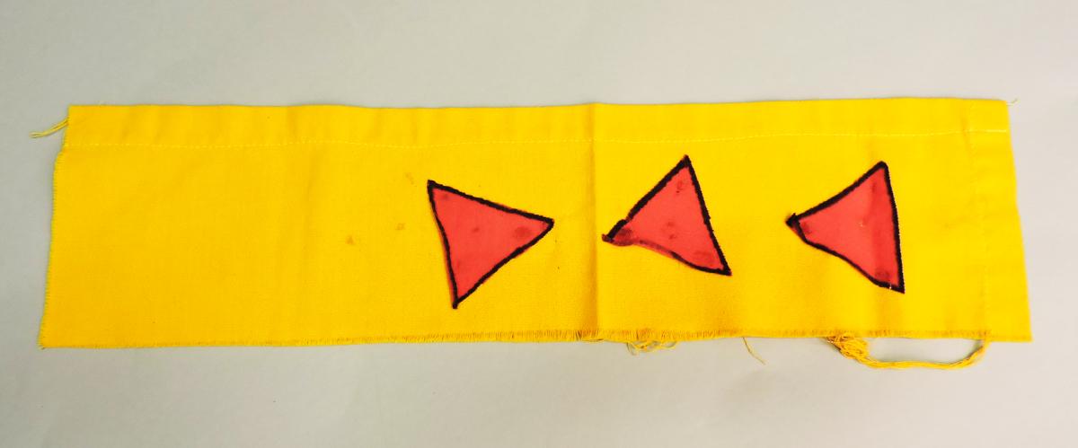 A roughly cut rectangular piece of yellow fabric. There is a seam on one long edge, the other edges are slightly frayed. On the fabric are three hand painted pink triangles with black outlines.
