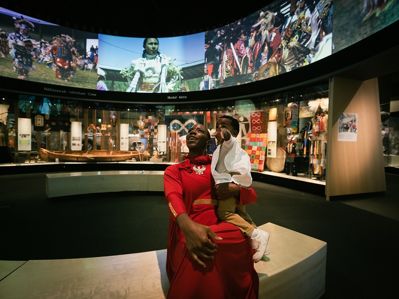 A woman and her young child sit in the circular What Makes Us Strong gallery, looking up in awe at the screens above.